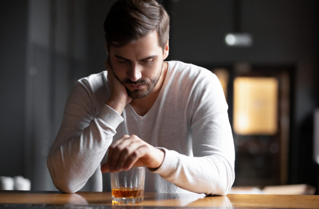 Upset young man sitting at bar alone with whisky drink trying to overcome addiction