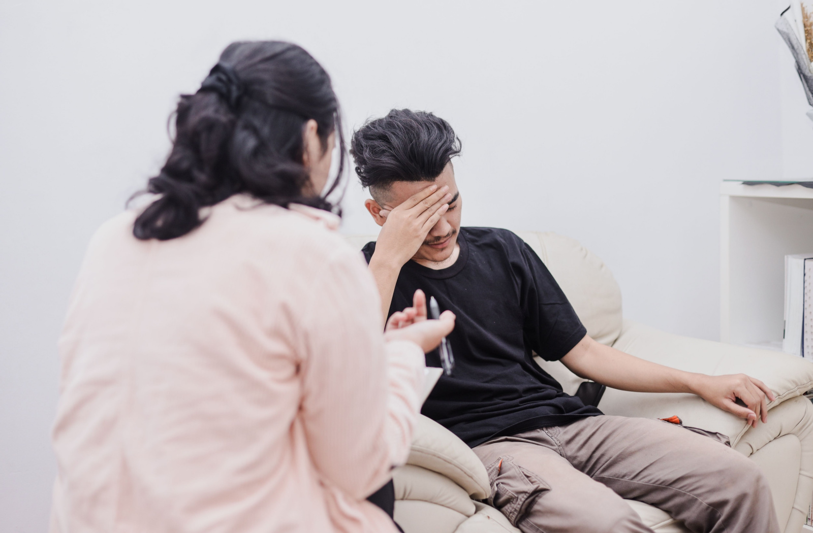 A young man in distress sitting on a sofa and listening to a psychologist while she gives him advice.