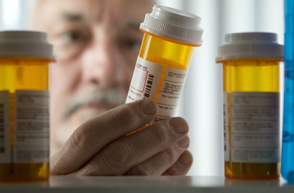 Close-up of 3 prescription pill bottles as a man holds one up to look at it. His face is out of focus in the background.